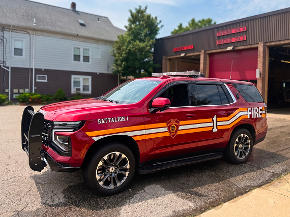 Red 'Battalion 1' fire command SUV with roof light bar and 'FIRE' decal parked outside a fire station bay, brown two-story building and open garage door with 'Engine Co. 3 / Rescue Co. 2' signage in the background.
